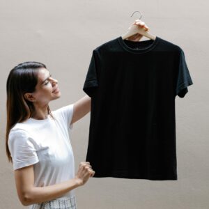 Side view of self assured young woman with long dark hair in stylish outfit smiling while holding hanger with black t shirt against gray wall